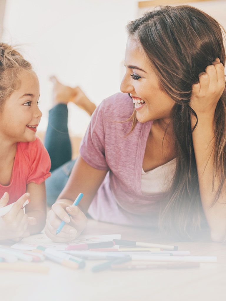 Eine Babysitterin liegt gemeinsam mit einem jungen Mädchen am Boden während sie mit vielen Buntstiften ein Bild malen und dabei herzlich lachen